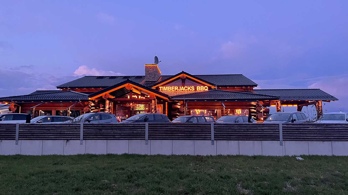 Timberjacks Bispingen Restaurant bei Dämmerung, rustikales Holzgebäude mit leuchtendem BBQ-Schild und geparkten Autos im Vordergrund.