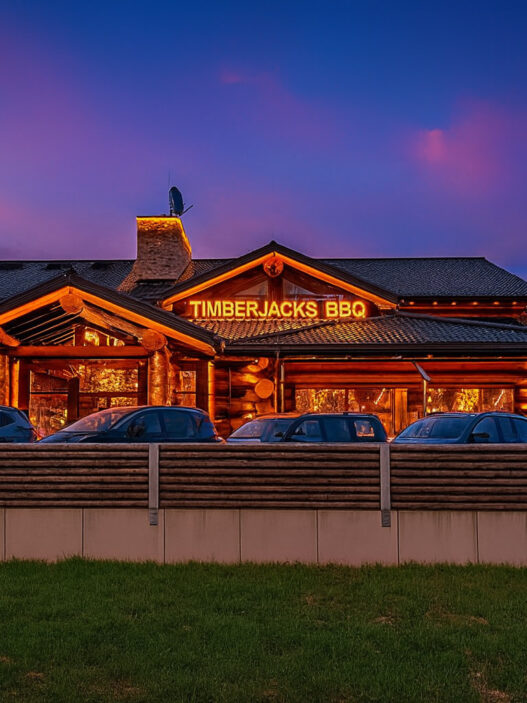 Timberjacks Bispingen Restaurant bei Dämmerung, rustikales Holzgebäude mit leuchtendem BBQ-Schild und geparkten Autos im Vordergrund.